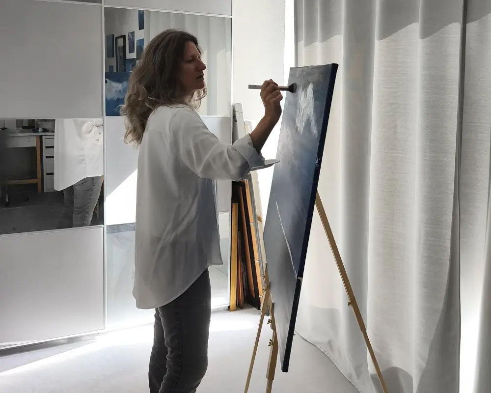 Une femme peint debout sur une toile posée sur un chevalet, dans un intérieur lumineux et moderne, pour illustrer la création artistique de Coralichanneling Art